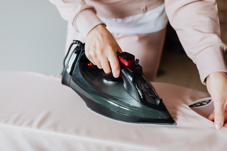 A person is using a modern black and red handheld steam iron to press a light-colored fabric surface, likely a bedsheet or cover, on a bed or sofa in a well-lit room. The scene focuses on the ironing 