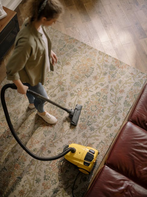 A professional cleaner dressed in a white protective suit, face mask, and gloves is operating a steam cleaning machine on a beige carpet in a modern residential living room. The room features natural