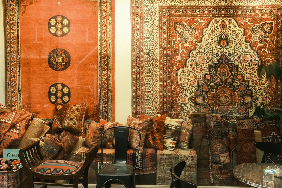 A neatly organized display of folded rugs and carpets in various patterns and colors, stacked on top of each other and around the room. The scene features different rug materials including wool and sy